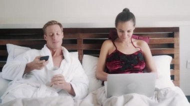 Aile çift yatakta yatıyor, erkek kahve içiyor, cep telefonu kullanıyor, kadın dizüstü bilgisayarla çalışıyor.