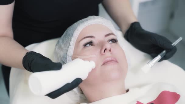 Przed zabiegiem kosmetyczka w rękawiczkach nakłada specjalny żel na twarz pacjenta — Wideo stockowe