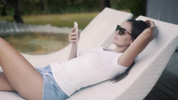 Tranquil woman uses smartphone lying on couch at resort — Stockvideo