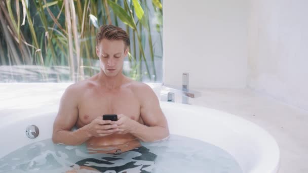 Young man reads messages on smartphone in modern bathtub — Stock Video