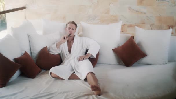 Blond guy in bathrobe switches on TV on comfortable bed — Αρχείο Βίντεο