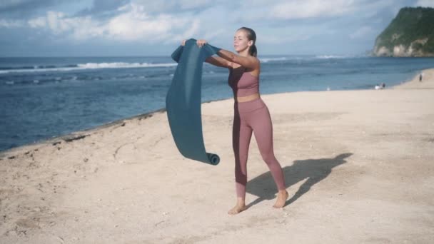 Blonde fille écarte tapis de yoga marine sur la plage de sable de l'océan — Video