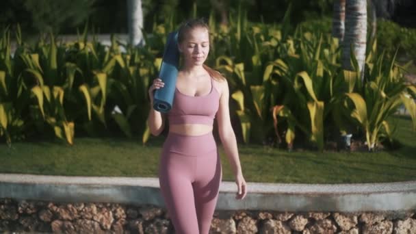 Menina em traje de treino caminha passado plantas tropicais segurando tapete — Vídeo de Stock