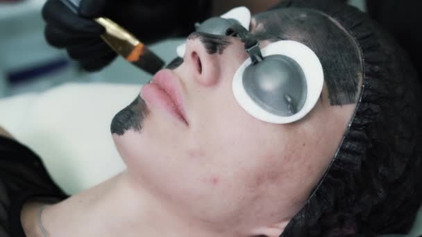 Close up, cosmetologist applies black carbon mask to skin of woman face — Stok video