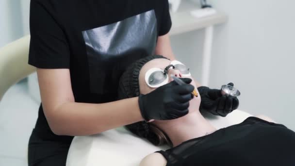 Крупным планом косметолог наносит черную маску на кожу женского лица — стоковое видео