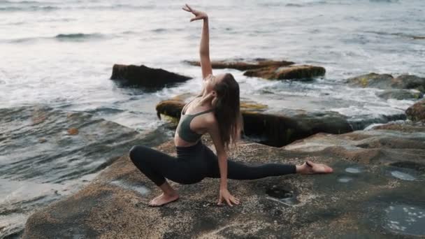 Chica hace estiramiento de pie en piedra contra agitar el océano al atardecer — Vídeos de Stock