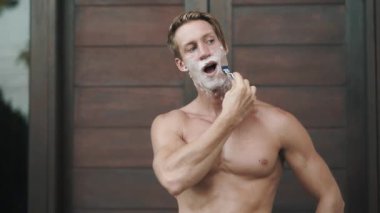 Portrait of man looking at camera and shaving his beard with manual razor