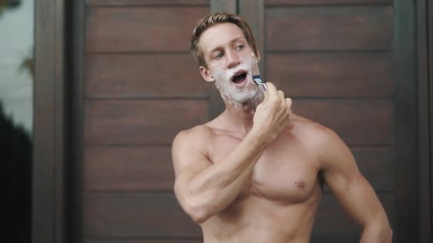 Portrait of man looking at camera and shaving his beard with manual razor — Stockvideo