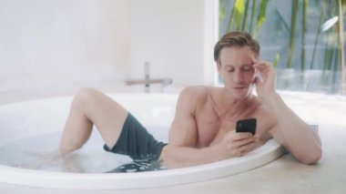 young man reads messages on smartphone in modern bathtub