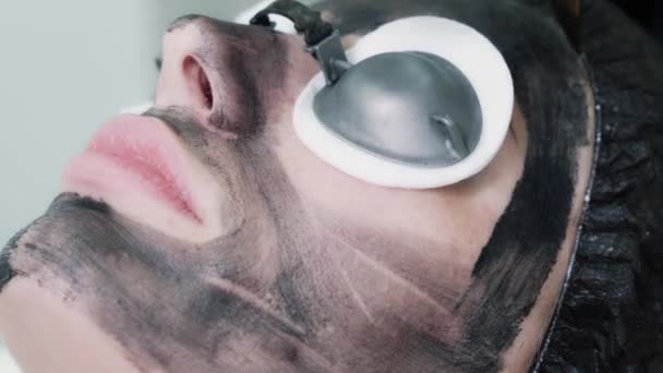 Κοντινό πλάνο, χέρι cosmetologist εφαρμόσει μαύρη μάσκα άνθρακα στο δέρμα της γυναίκας πρόσωπο — Αρχείο Βίντεο