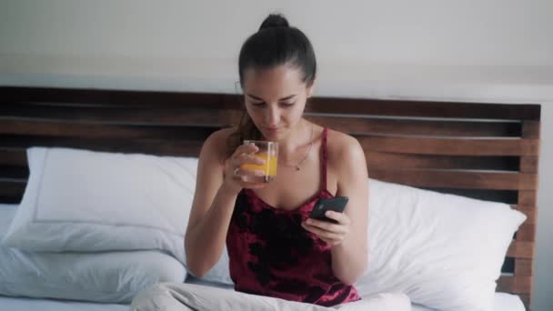 Hermosa chica con vaso de jugo navega por Internet con teléfono inteligente en la cama grande — Vídeo de stock