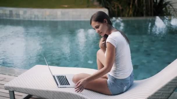 Seitenansicht des fröhlichen Mädchens arbeitet am Laptop sitzt auf der Couch in der Nähe des Pools — Stockvideo