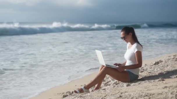 Mulher de óculos de sol trabalha na praia com laptop, abre e começa a digitar — Vídeo de Stock