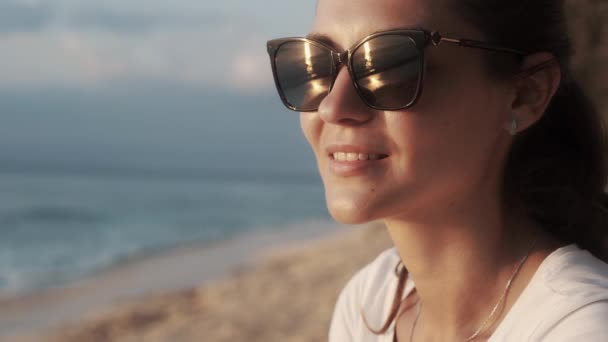 Porträt einer schönen lächelnden Frau mit Sonnenbrille, die das Meer bei Sonnenuntergang betrachtet. — Stockvideo