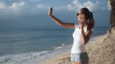 Güneş gözlüklü kadın gün batımında deniz kenarında akıllı telefondan selfie çekiyor..
