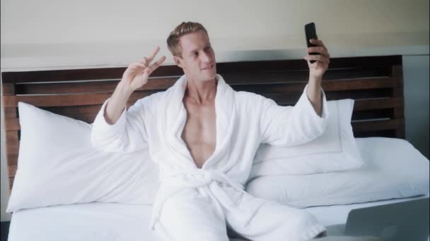 Beau blond mec en peignoir blanc faire selfie photo sur téléphone mobile dans le lit — Video