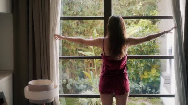 Zadní pohled žena v pyžamu rozepíná závěsy v moderní ložnici, zpomaluje — Stock video