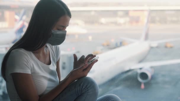 Женщина в защитной маске пользуется смартфоном, отправляет сообщение в аэропорт COVID-19 — стоковое видео