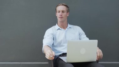 Adam modern dizüstü bilgisayarla oturur ve arka planda gri bir duvar ile sigara içer.