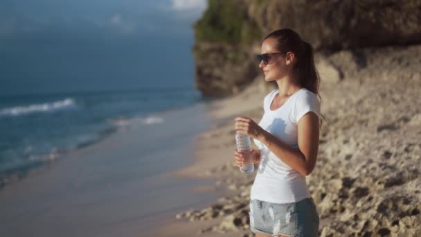 Krásná mladá žena v slunečních brýlích pije čistou vodu z láhve na pláži — Stock video