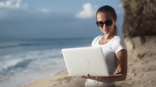 Freelancer mulher em óculos de sol detém laptop, tipos e obras na praia tropical — Vídeo de Stock