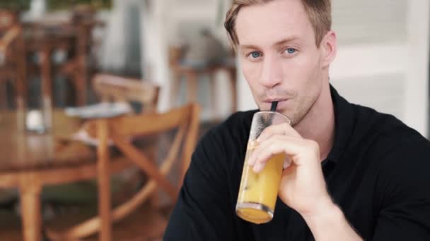 Retrato del hombre guapo bebiendo jugo de naranja en la cafetería, mira a la cámara, sonríe — Vídeo de stock