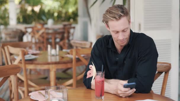 Hombre guapo con camisa se sienta en la cafetería, bebe jugo y usa el teléfono — Vídeos de Stock