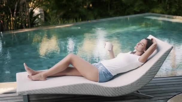 Mujer joven en ropa casual se encuentra en la cama de sol cerca de la piscina y bebe café — Vídeos de Stock