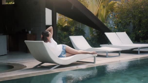 Side view, woman in T-shirt and shorts lies on sunbed near pool and rests — Stock Video