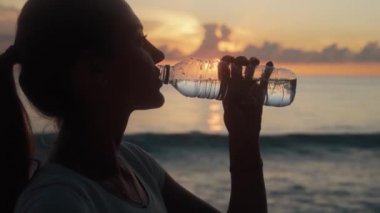 Kadın silueti, okyanus sahilinde gün batımında şişeden temiz su içer.