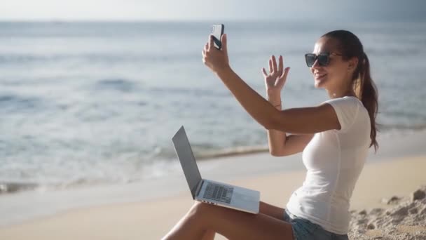 Женщина с ноутбуком сидит на пляже возле океана и использует телефон для видеозвонка — стоковое видео