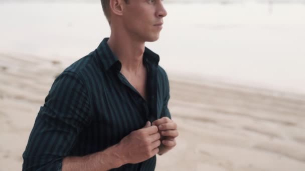 검은 셔츠를 입고 모래 해변을 거닐고 있는 잘생긴 남자의 모습. — 비디오