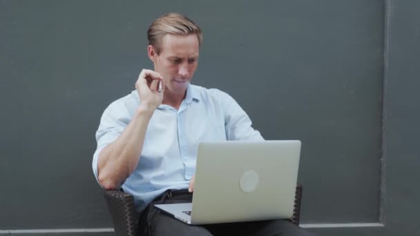 Homem fuma nervosamente cigarro, olha para a tela do laptop, parede cinza no fundo — Vídeo de Stock