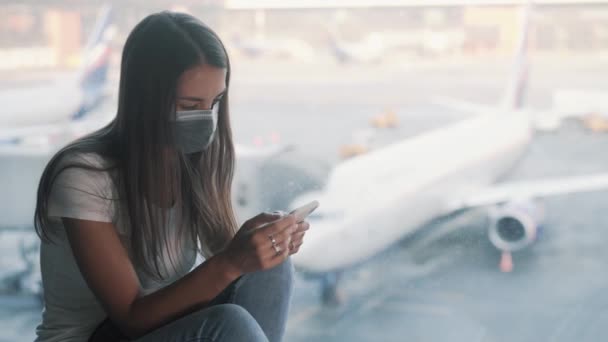 Mulher em máscara protetora senta perto da janela no aeroporto, usa smartphone COVID-19 — Vídeo de Stock