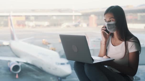 Meisje met beschermend masker maakt gebruik van laptop en gesprekken op de telefoon op de luchthaven, COVID-19 — Stockvideo