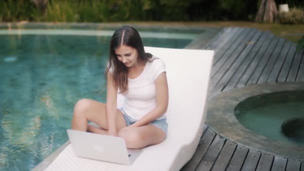 Женщина-фрилансер сидит на шезлонге возле бассейна, работает на ноутбуке, получает хорошие новости — стоковое видео