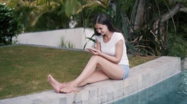 Genç bir kadın havuzun kenarında oturuyor ve iş için telefon kullanıyor.
