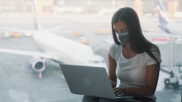保護マスクの女性は空港の窓の近くに座っており、ラップトップを使用しています, COVID-19 — ストック動画