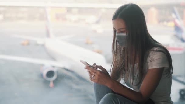 Donna in maschera protettiva si siede vicino alla finestra in aeroporto, utilizza smartphone COVID-19 — Video Stock