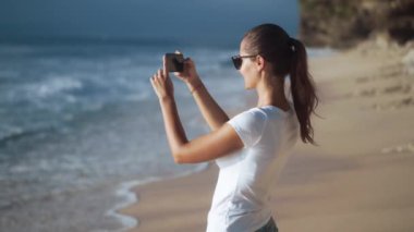 Güneş gözlüklü neşeli genç bir kadın plajın fotoğraflarını çekiyor.