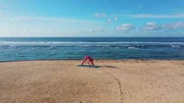 Drone görüntüleri. Kadın, okyanus manzaralı plajda yoga egzersizleri yapıyor ve esniyor. 