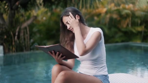 Mujer joven en ropa de verano se sienta en la cama de sol y lee libro cerca de la piscina — Vídeo de stock