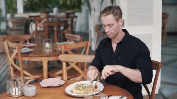El hombre se sienta a la mesa en la cafetería y come comida, la corta con cuchillo y tenedor en el plato — Vídeos de Stock