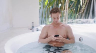 Genç adam lüks dairelerde modern yuvarlak küvette otururken akıllı telefon kullanıyor.