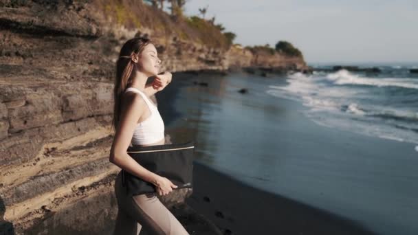 Dziewczyna w odzieży sportowej pozuje przed kamerą na plaży przed falami oceanu, zwolnione tempo — Wideo stockowe