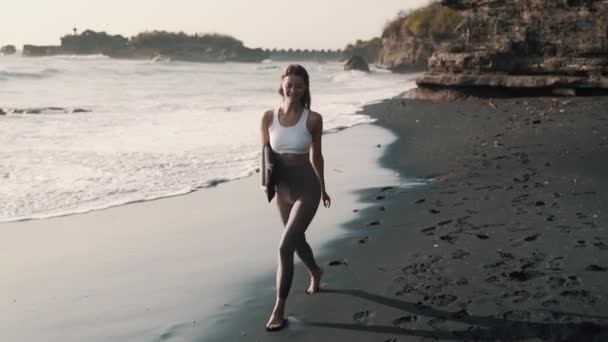 Happy woman in sportswear walks along black sand beach, smiles, turns around — Stock Video