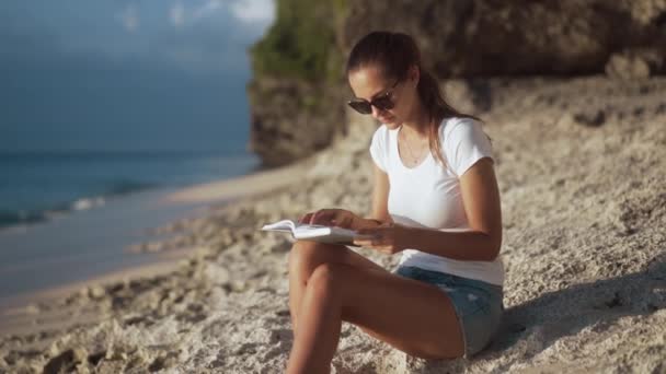 Очаровательная девушка читает книгу, сидя на песчаном берегу — стоковое видео