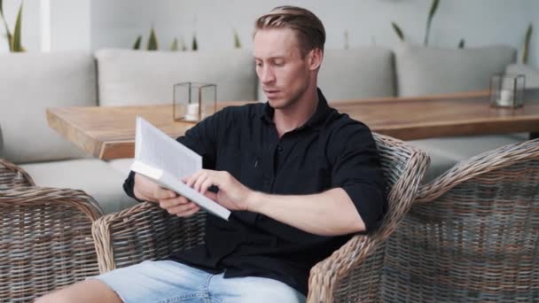 Mann sitzt im Korbsessel aus Rattan und liest interessantes Buch in modernem Café — Stockvideo