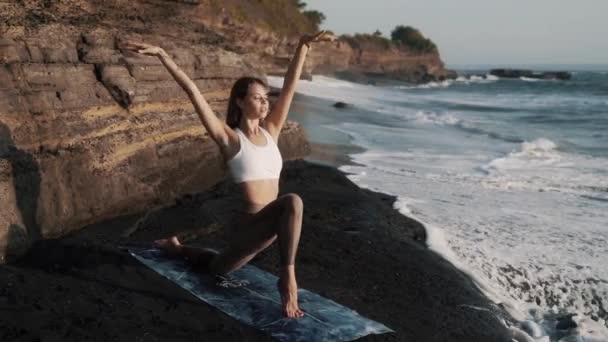 Дівчина розтягується на килимку на кам'яному чорному пляжі біля океану — стокове відео
