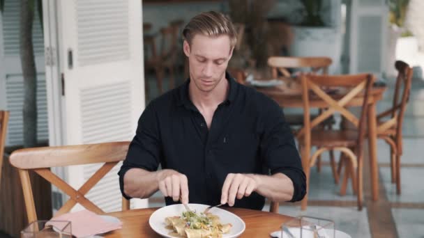 Mannen sitter vid bordet på caféet och skär måltid med kniv och gaffel i tallriken — Stockvideo